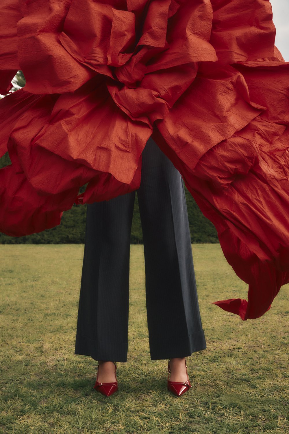 Fashion shot featuring a person wearing dark trousers and red heels, standing on grass, framed by a large red textured fabric.