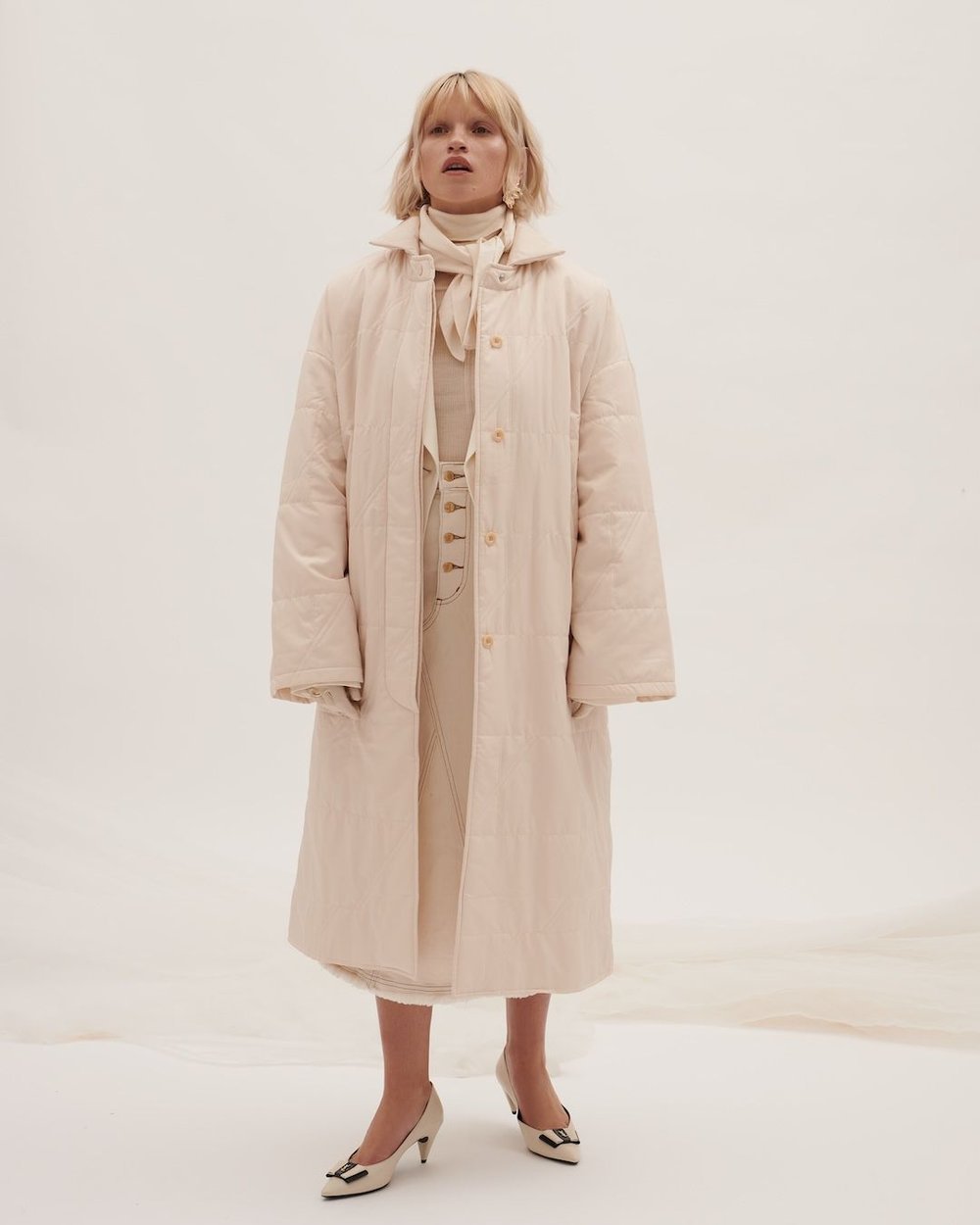 A woman stands in a white environment wearing a quilted coat, light-colored dress, pumps, and a neck scarf with natural lighting.
