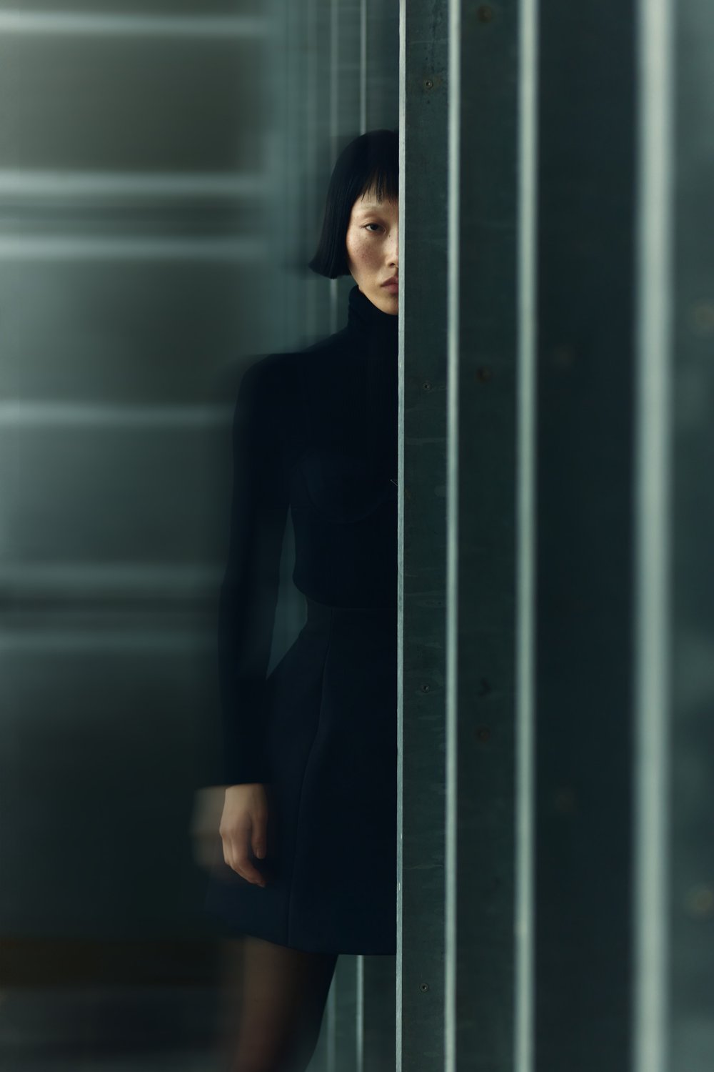 Portrait of a stylish Asian woman in a black turtleneck dress posing indoors between vertical grey metal bars, artistic fashion photography.