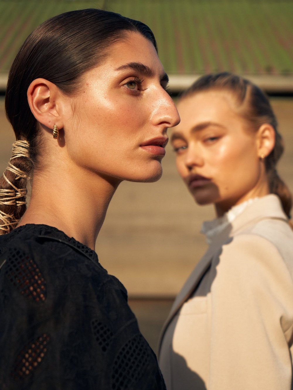 Two women with sleek dark and blonde hair, wearing black and beige jackets respectively, pose in an outdoor setting during daytime.