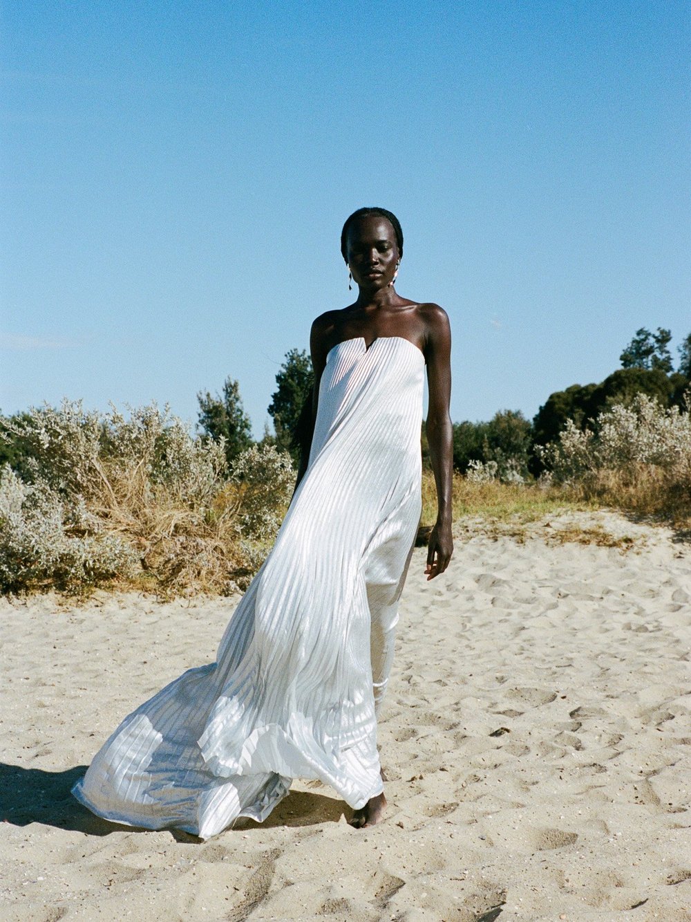 A woman in a white gown poses on a sandy beach with dunes and vegetation under a clear blue sky in an outdoor fashion shot.