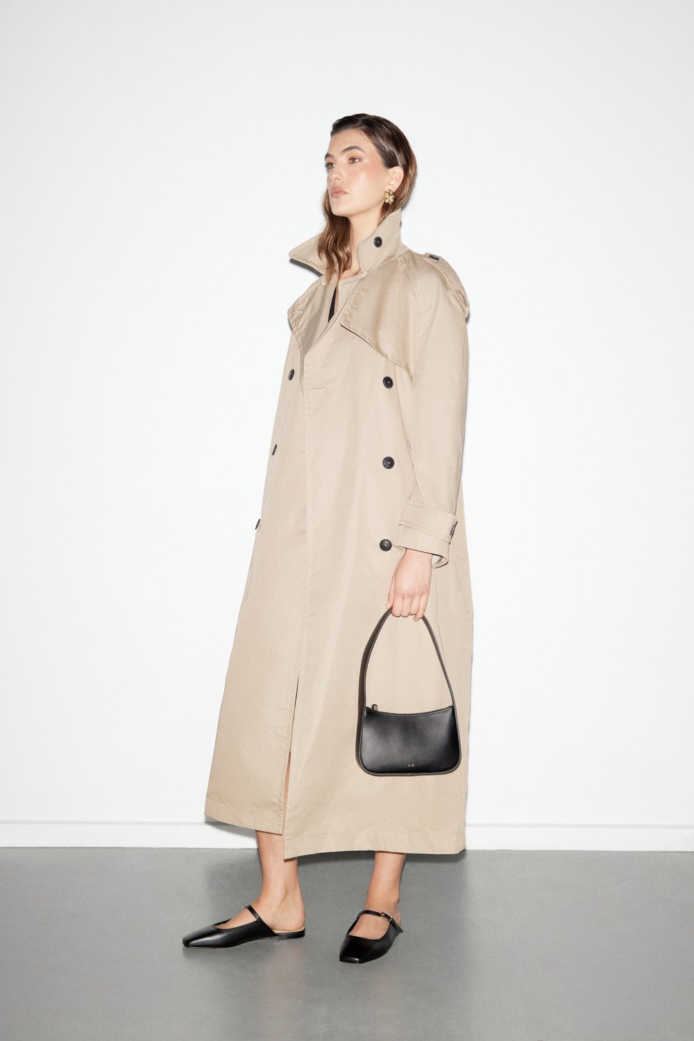Woman in trench coat with black purse and flats stands in a white studio, showcasing a fashion-forward and minimalist aesthetic.