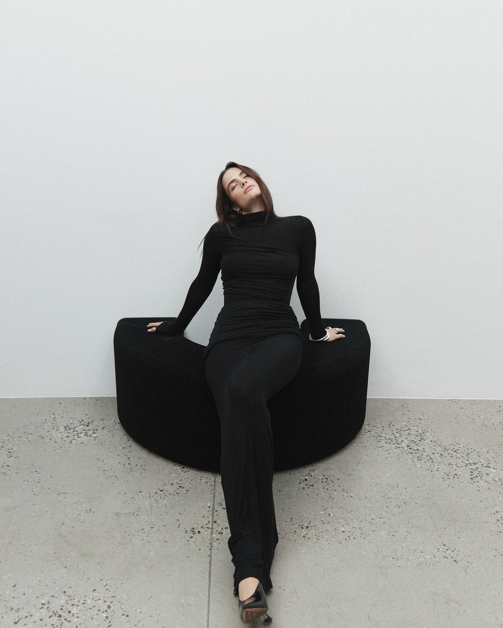 A woman in a black turtleneck dress poses on a curved black sofa against a white wall and gray floor, looking up with her eyes closed.