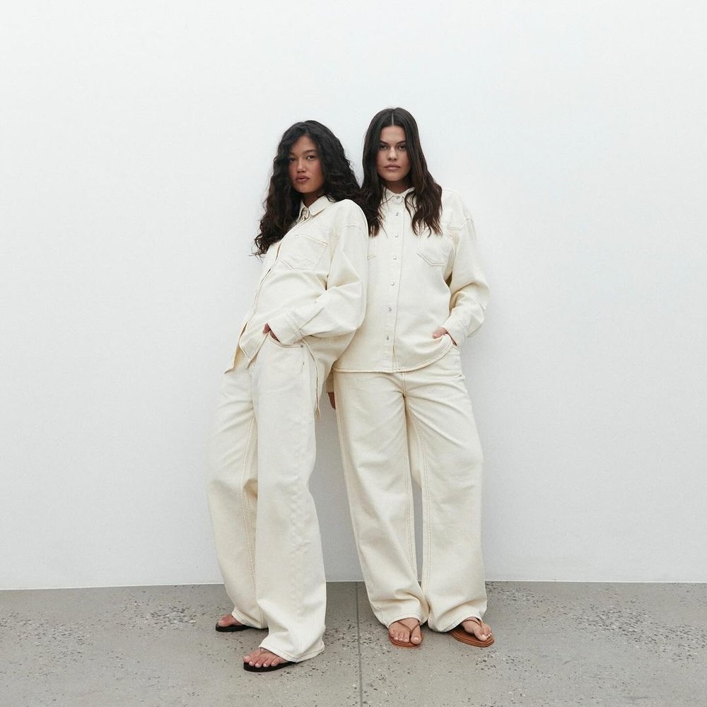 Two women in matching cream-colored outfits stand closely, leaning against a white wall, creating a minimalist fashion portrait.