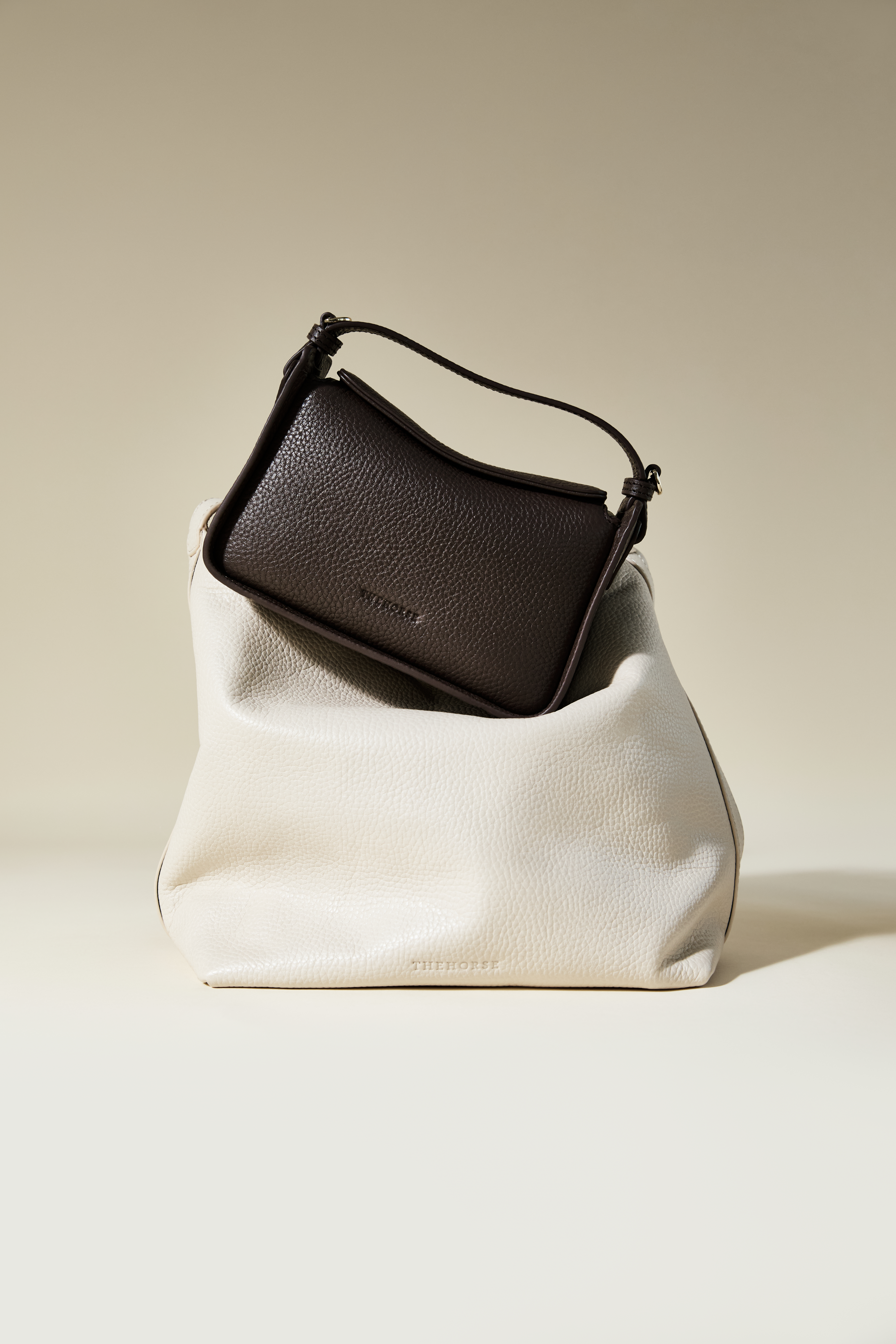 Two textured handbags in a studio setting. A small dark brown handbag is nested in a larger, cream-colored handbag on a beige surface.