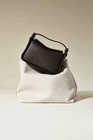 Two textured handbags in a studio setting. A small dark brown handbag is nested in a larger, cream-colored handbag on a beige surface.