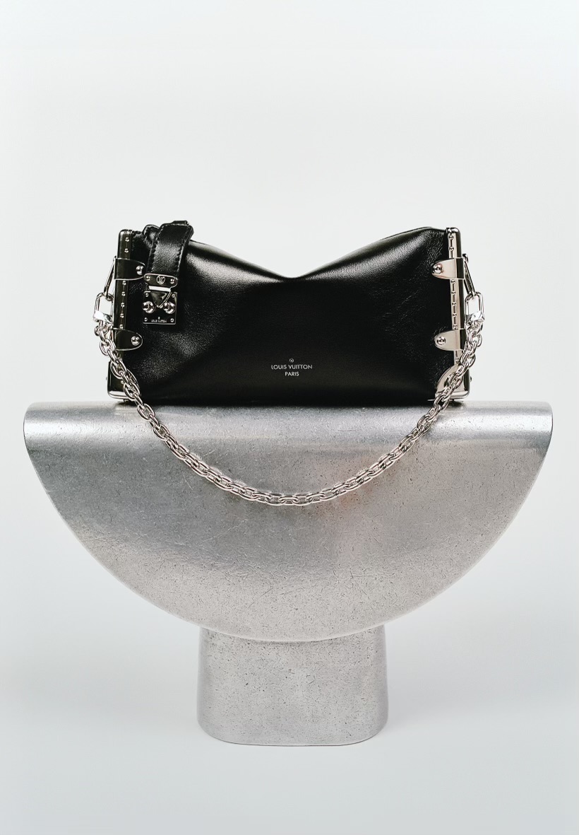 Black leather Louis Vuitton handbag with metal hardware and silver chain strap displayed on a silver metal stand against a white backdrop.