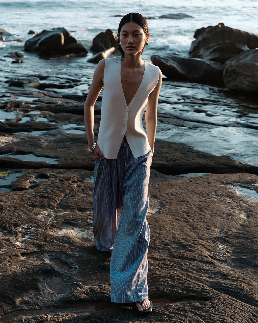 An Asian woman poses on a rocky beach in a sleeveless top and wide-leg striped pants, with the ocean and sky as a backdrop. 