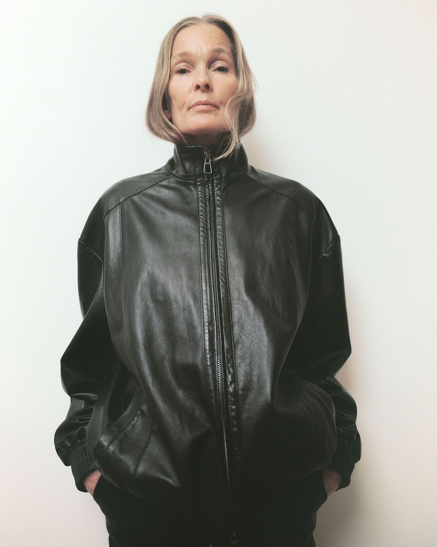 An adult woman poses indoors wearing a black leather jacket and black pants against a plain white backdrop in a medium studio shot.