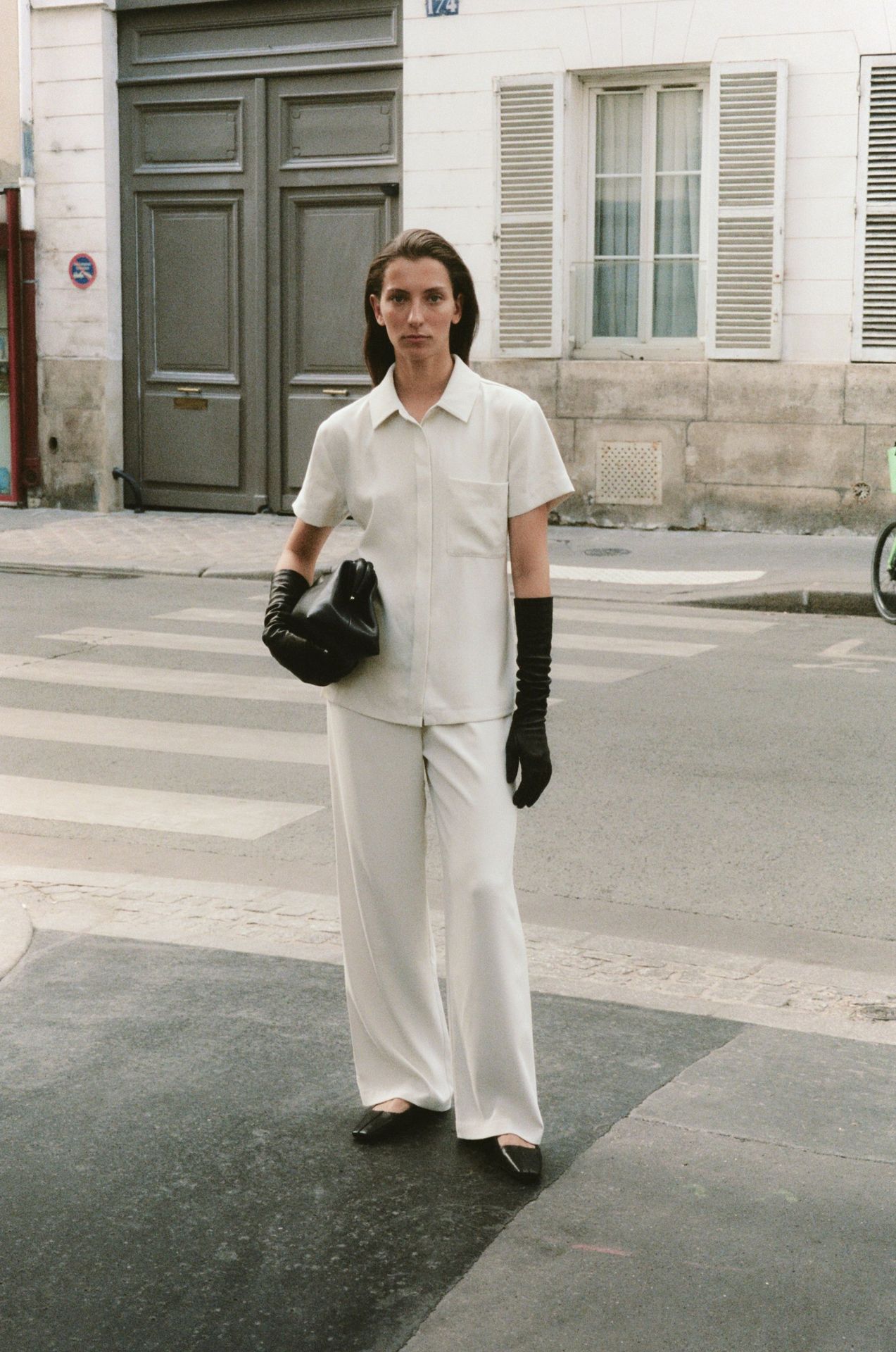 Adult female wearing a white outfit and black gloves stands on a crosswalk in an urban setting during daytime, holding a black bag.