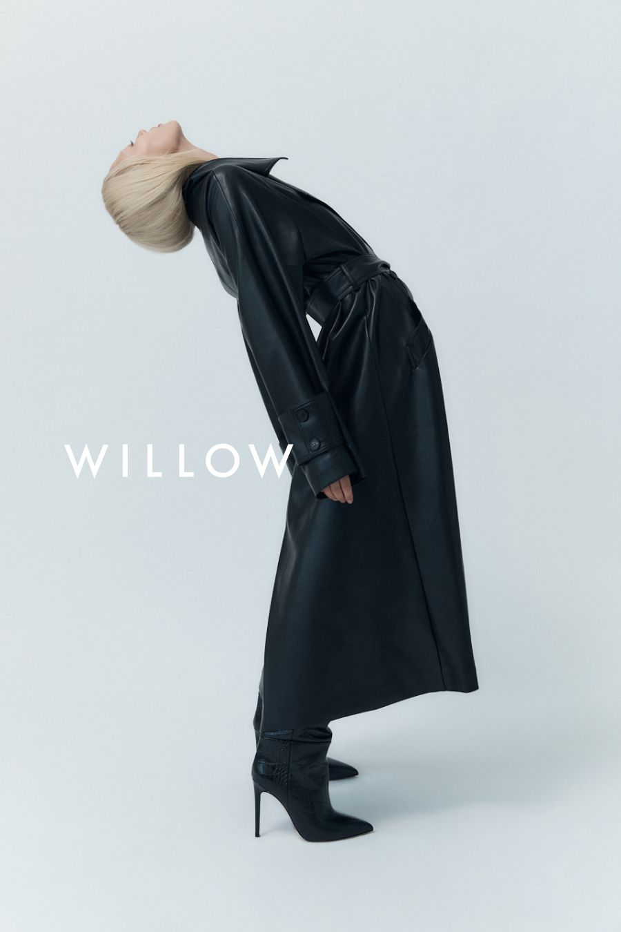 Fashion shot of a slender blonde woman in a long black leather coat and high-heeled boots, bending backward in a white studio.