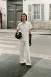 A woman wearing a cream button-down shirt and matching trousers stands on a city street holding a black leather handbag.