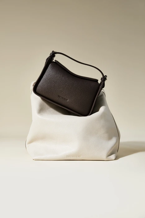 Dark brown leather handbag resting on a larger cream-colored slouchy bag against a neutral background.