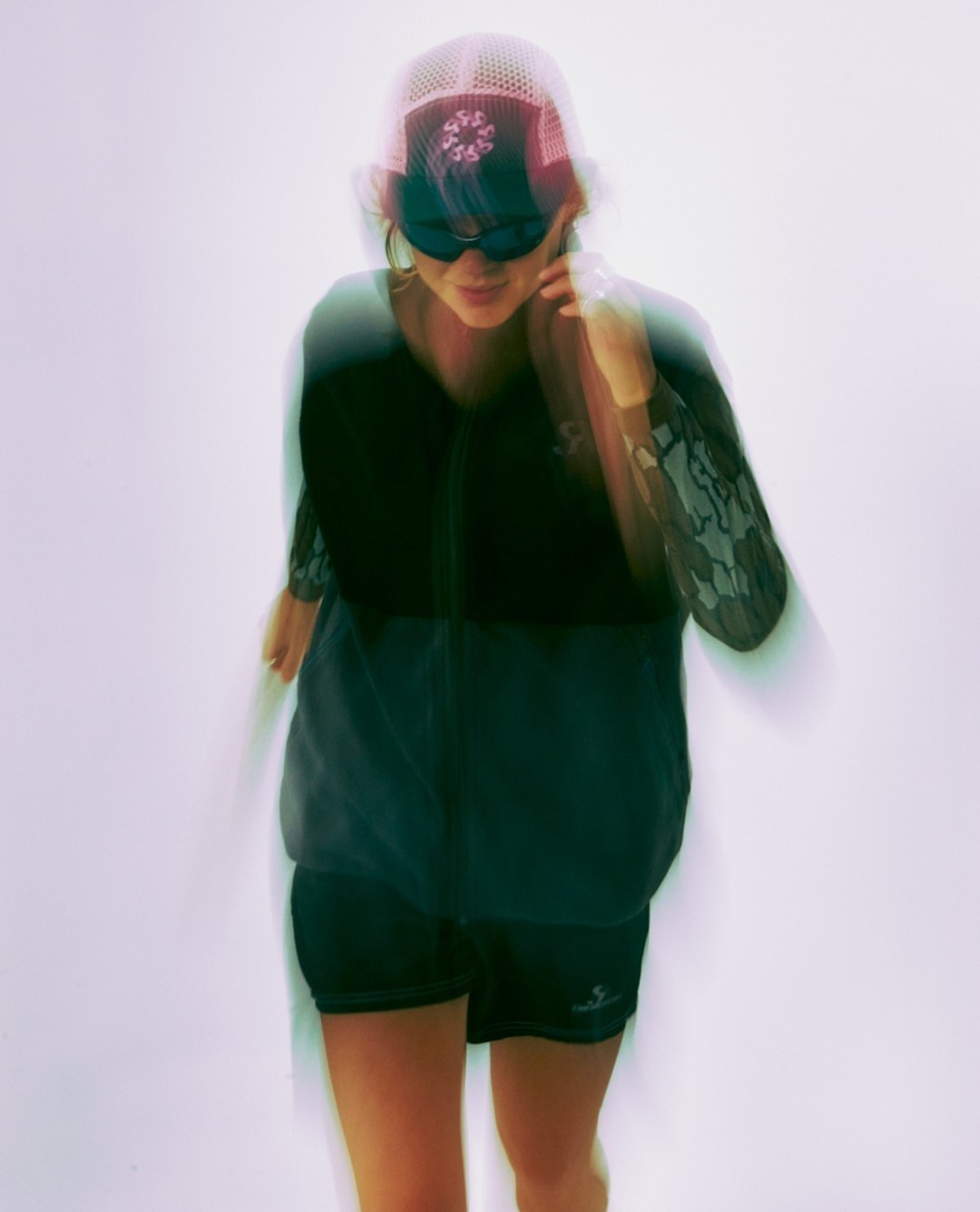 Woman in sunglasses and athletic wear in motion against a white background with long exposure creates a blurred effect in a studio setting.