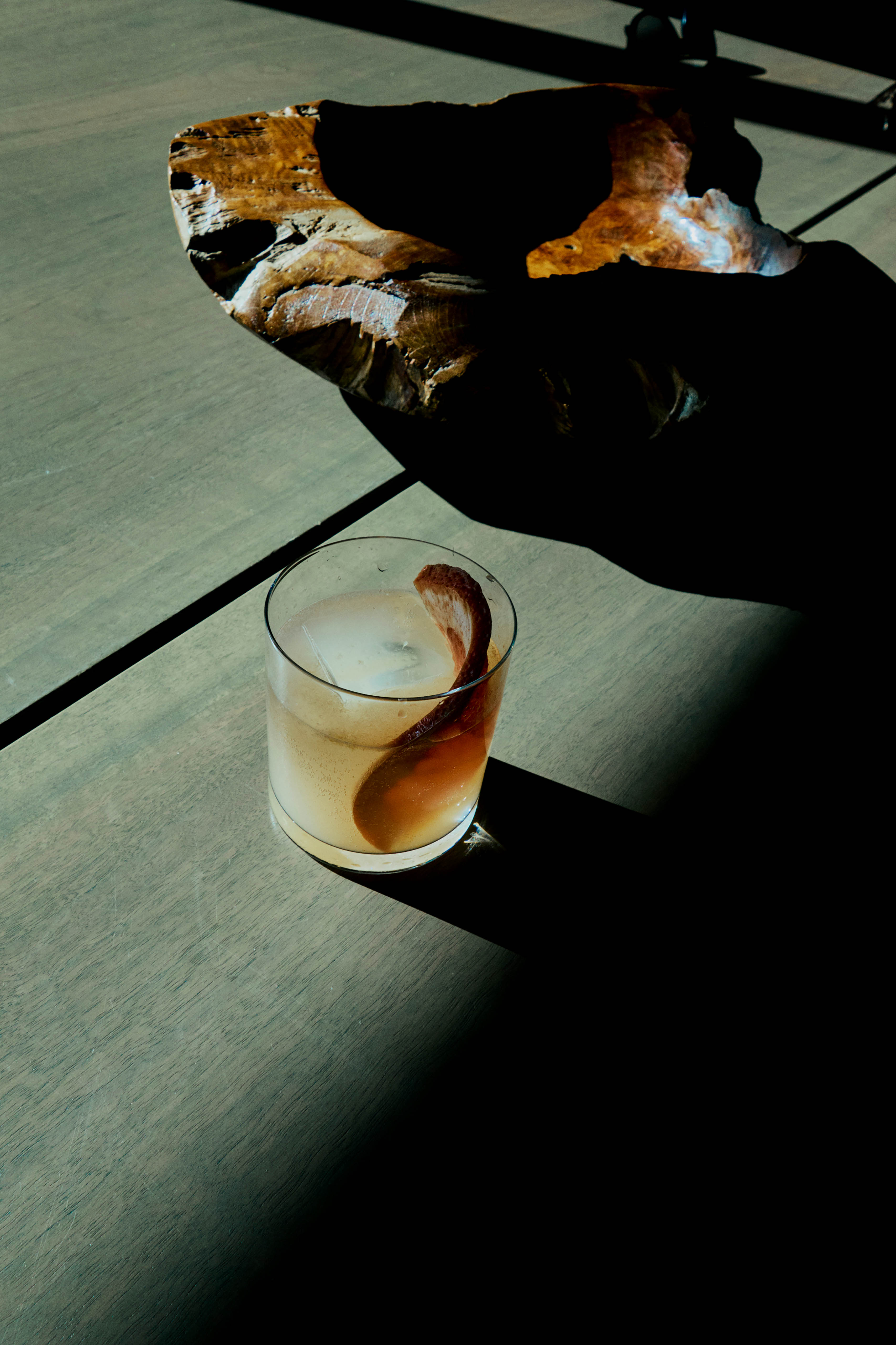 A cocktail in a glass with a large ice cube and orange peel twist, set on a table next to a geological rock formation under bright light.