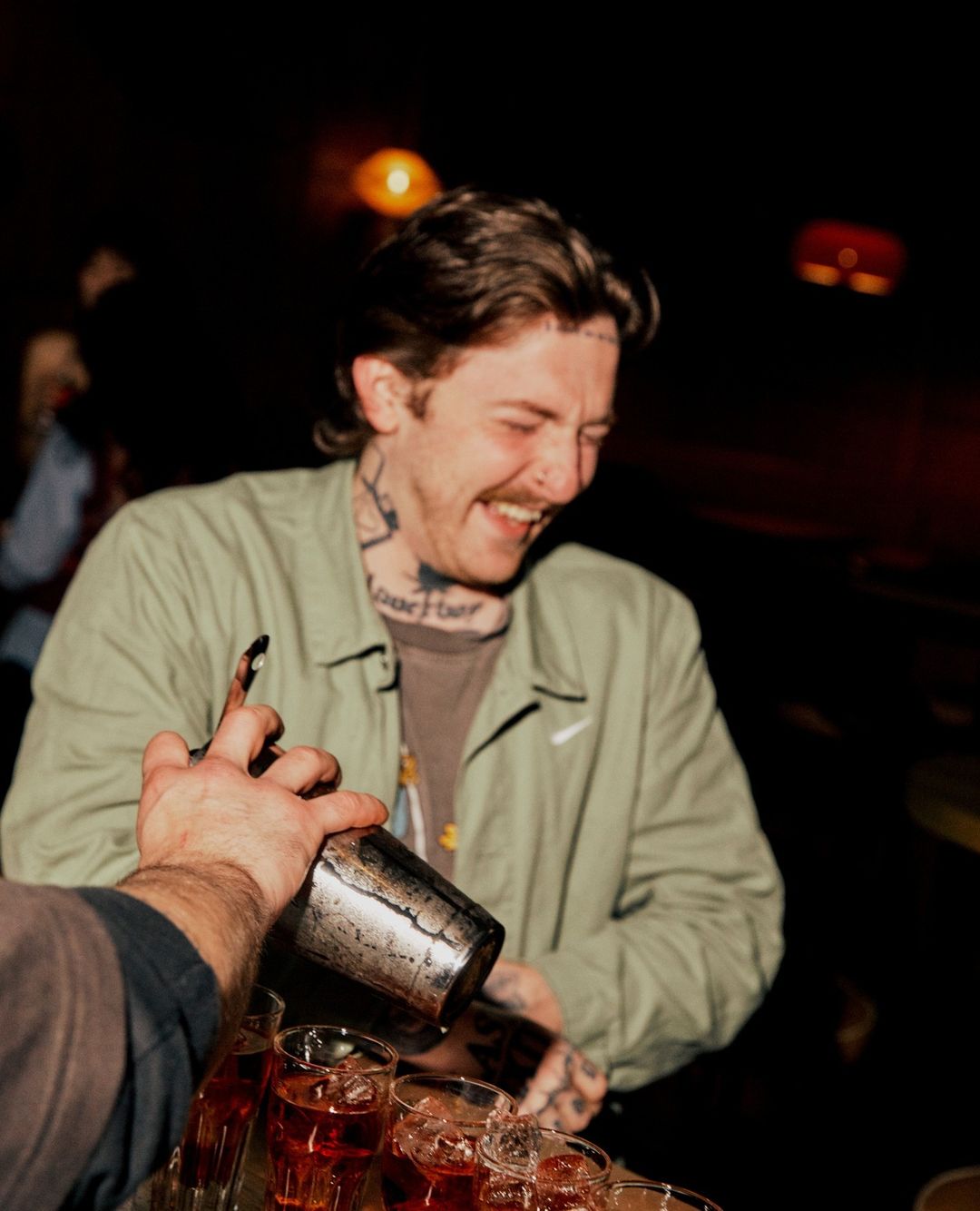 Bartender pouring cocktail into champagne coupes. Tattoos are visible on neck and hands. Light skin male smiling in dimly lit bar.