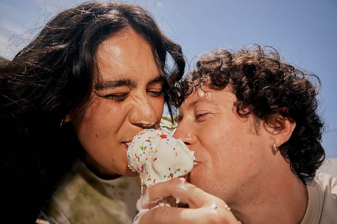 Close-up of a couple eating an ice cream cone with sprinkles under a bright blue sky, faces covered in melting ice cream.