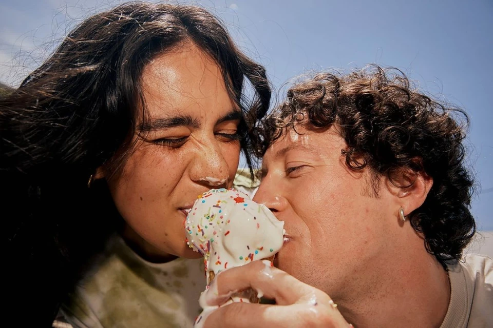 Close-up of a couple eating an ice cream cone with sprinkles under a bright blue sky, faces covered in melting ice cream.