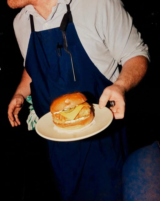 Chef in a blue apron presents a gourmet burger on a white plate in a dimly lit setting, highlighting food presentation and culinary expertise.
