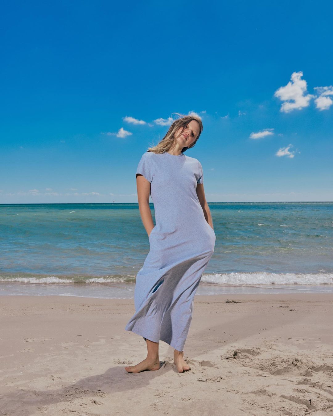 Young woman in light blue dress posing on a sandy beach under a blue sky with clouds and ocean in the background, enjoying the sunny weather.