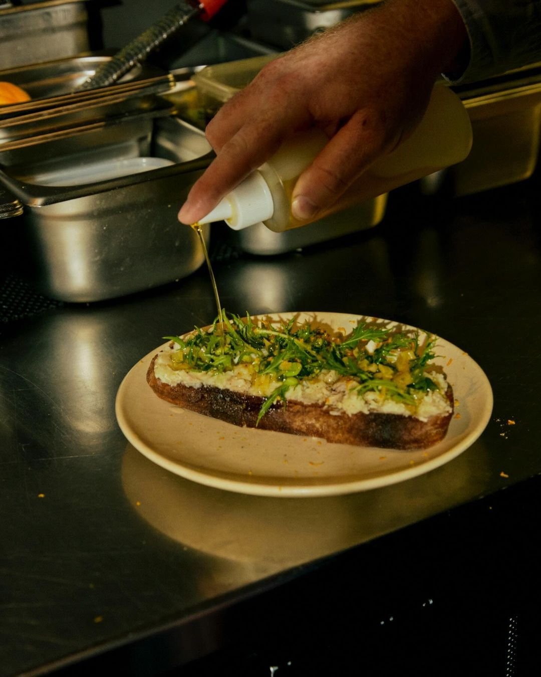 A food preparation scene shows a hand drizzling oil from a bottle onto a toast topped with greens and cheese on a plate, indoor kitchen.
