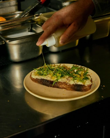 A food preparation scene shows a hand drizzling oil from a bottle onto a toast topped with greens and cheese on a plate, indoor kitchen.