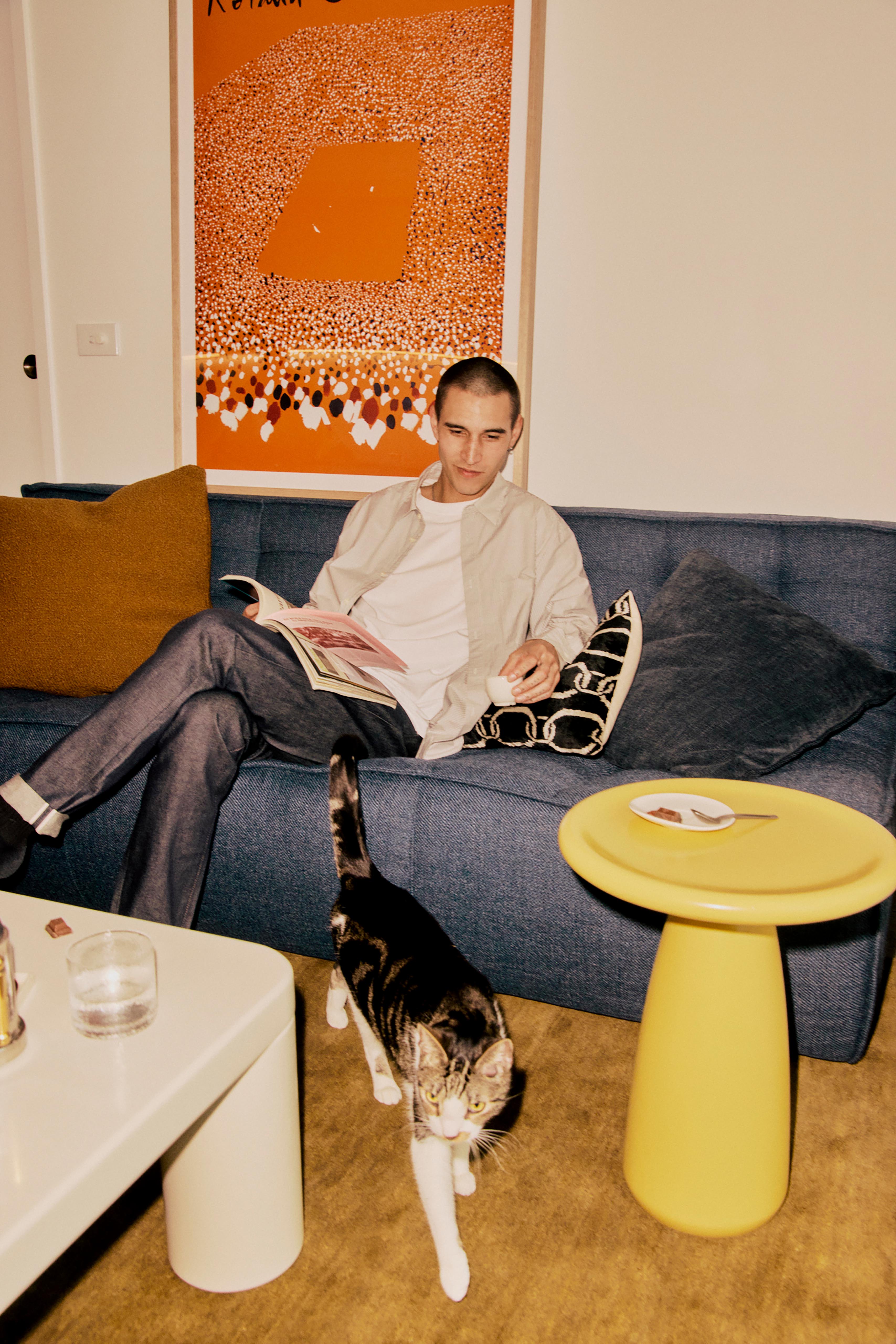 Man sits on a blue couch in a living room reading a magazine while a tabby cat walks by a yellow side table and white coffee table.