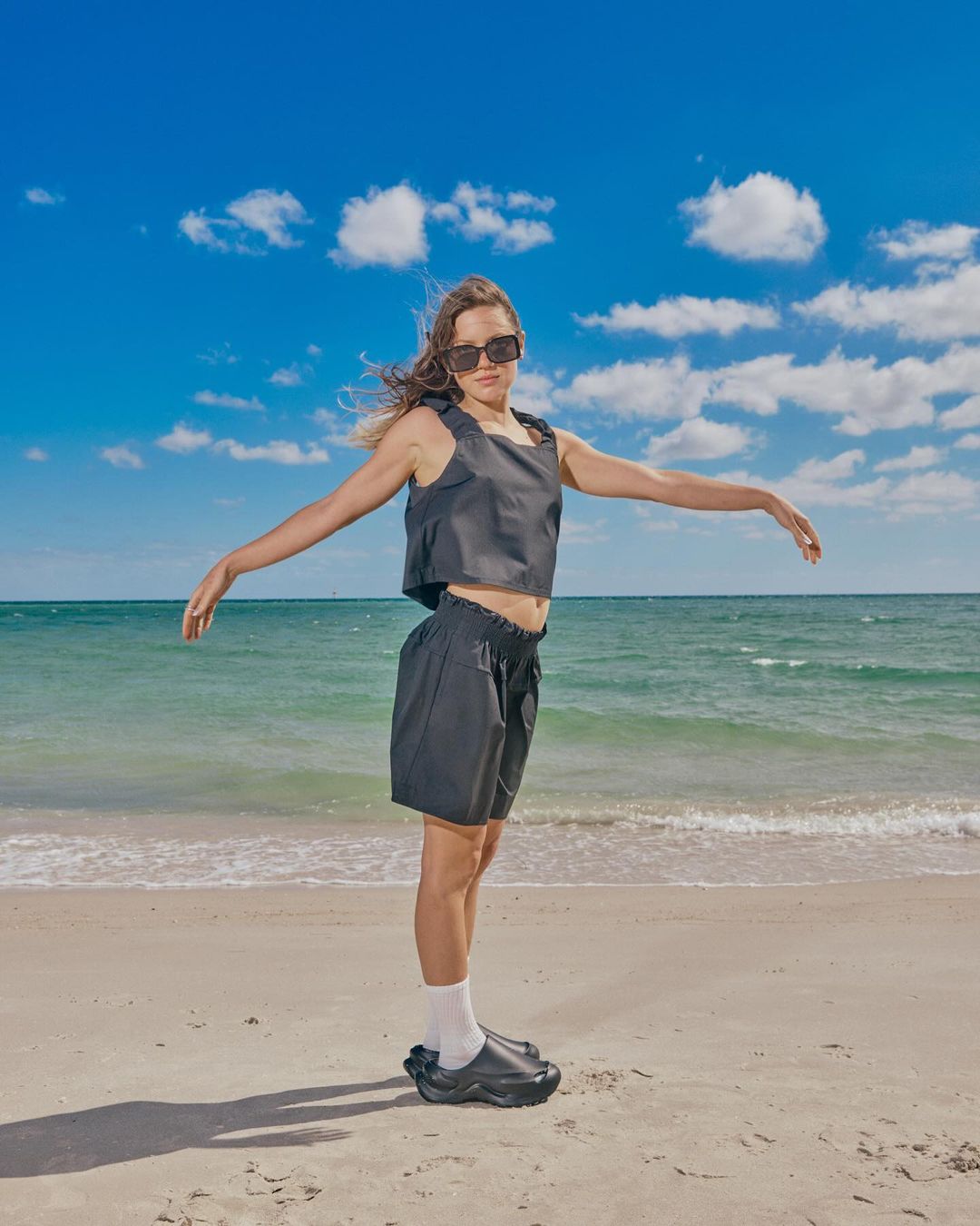 Woman in dark outfit poses on a sandy beach under a bright blue sky with clouds. Ocean waves are visible in the background.