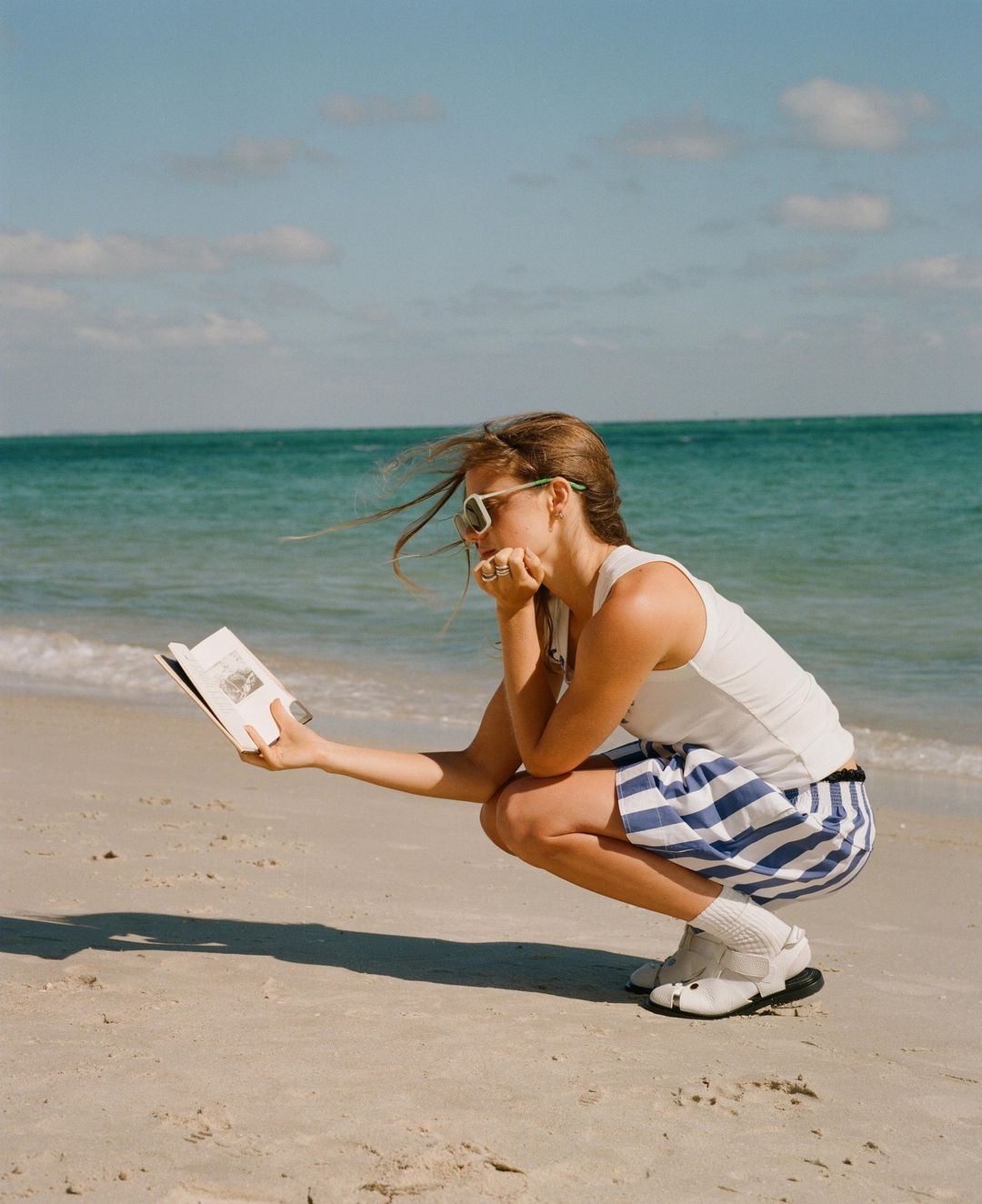 A woman in a white tank top and blue striped shorts squats on a sandy beach, holding open a book while the ocean water ripples behind her.