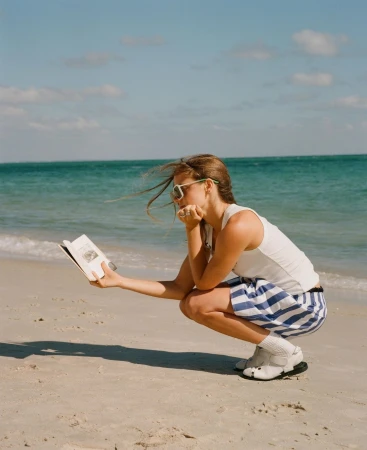 A woman in a white tank top and blue striped shorts squats on a sandy beach, holding open a book while the ocean water ripples behind her.
