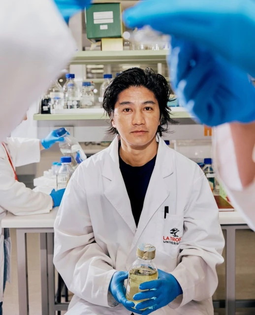 Asian scientist in a lab coat holds a bottle in a lab with other researchers working in the background, laboratory setting.