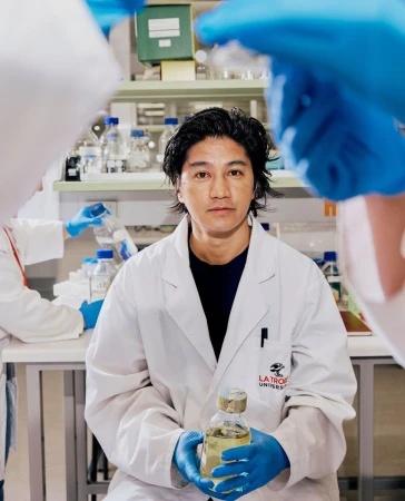 Asian scientist in a lab coat holds a bottle in a lab with other researchers working in the background, laboratory setting.