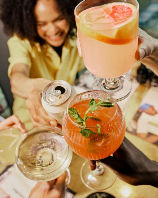 Group of friends clinking glasses and drinks including cocktails and a can together at a restaurant or bar, celebrating a fun night.