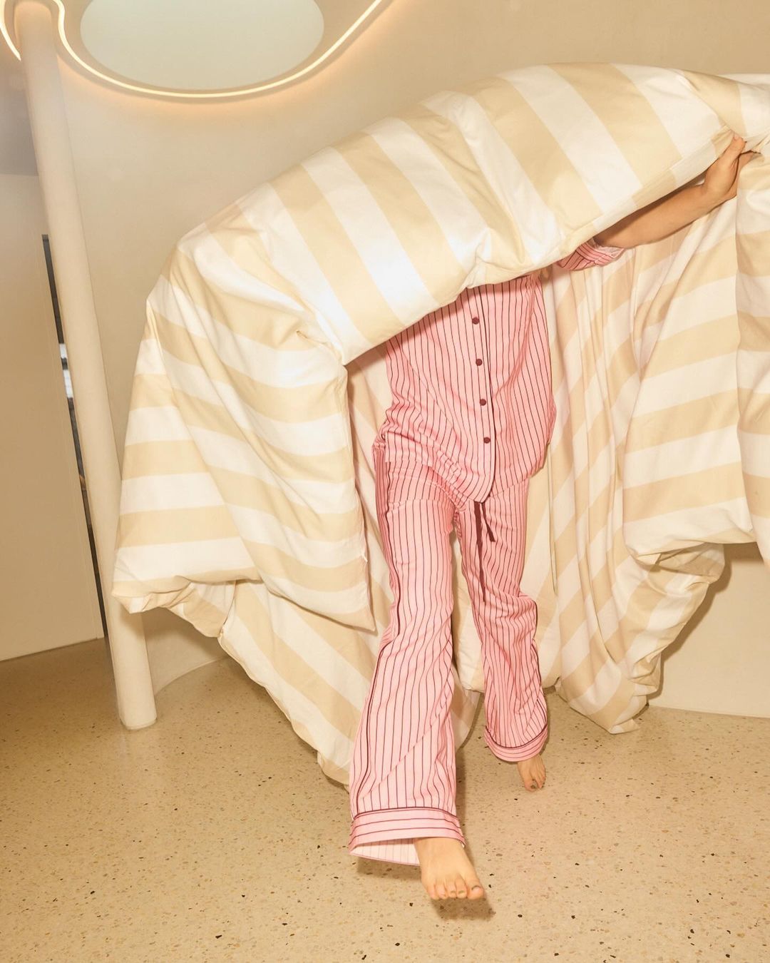 A person in striped pink pajamas stands behind a striped beige and white duvet in a brightly lit interior space. 