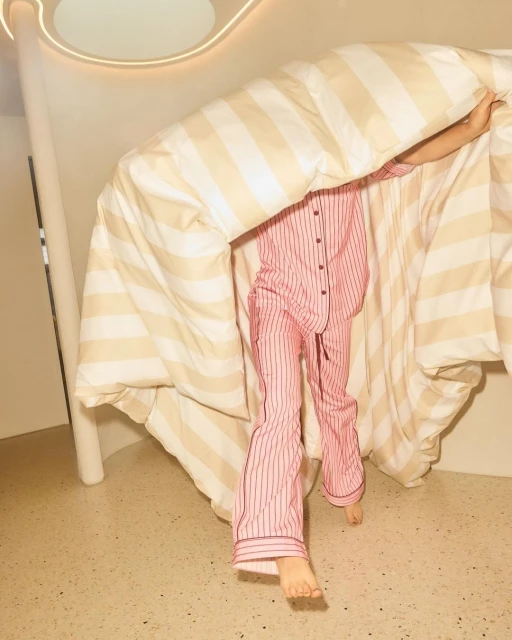 A person in striped pink pajamas stands behind a striped beige and white duvet in a brightly lit interior space. 