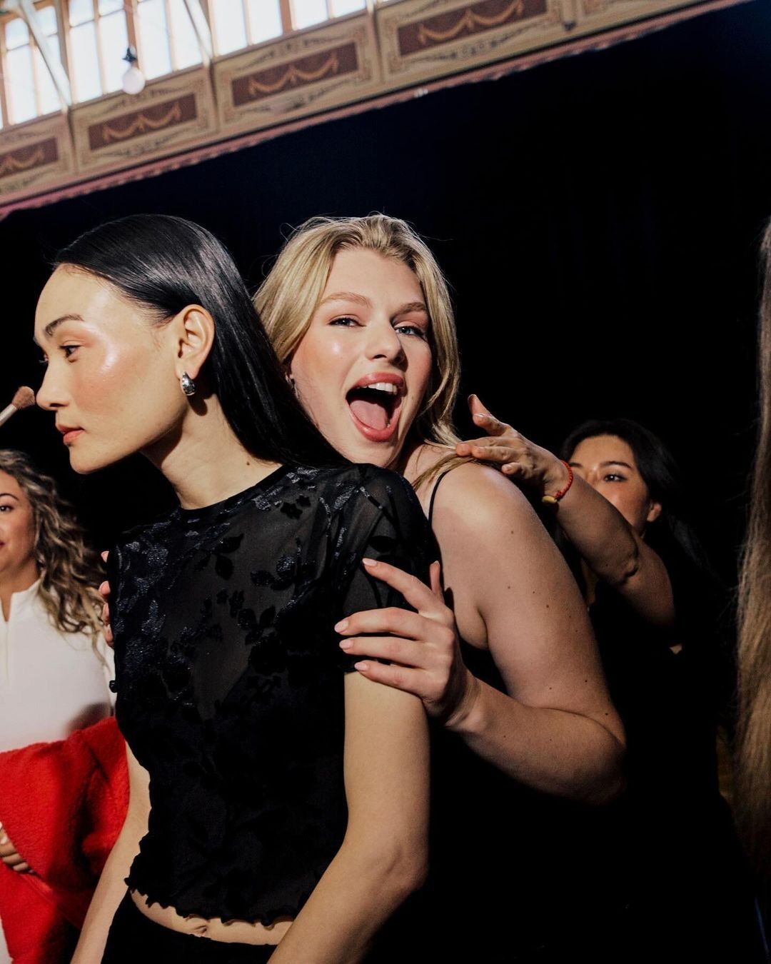 Two adult women in a dark indoor setting, one blonde with mouth open, the other with black hair in black mesh top, with others in the background.