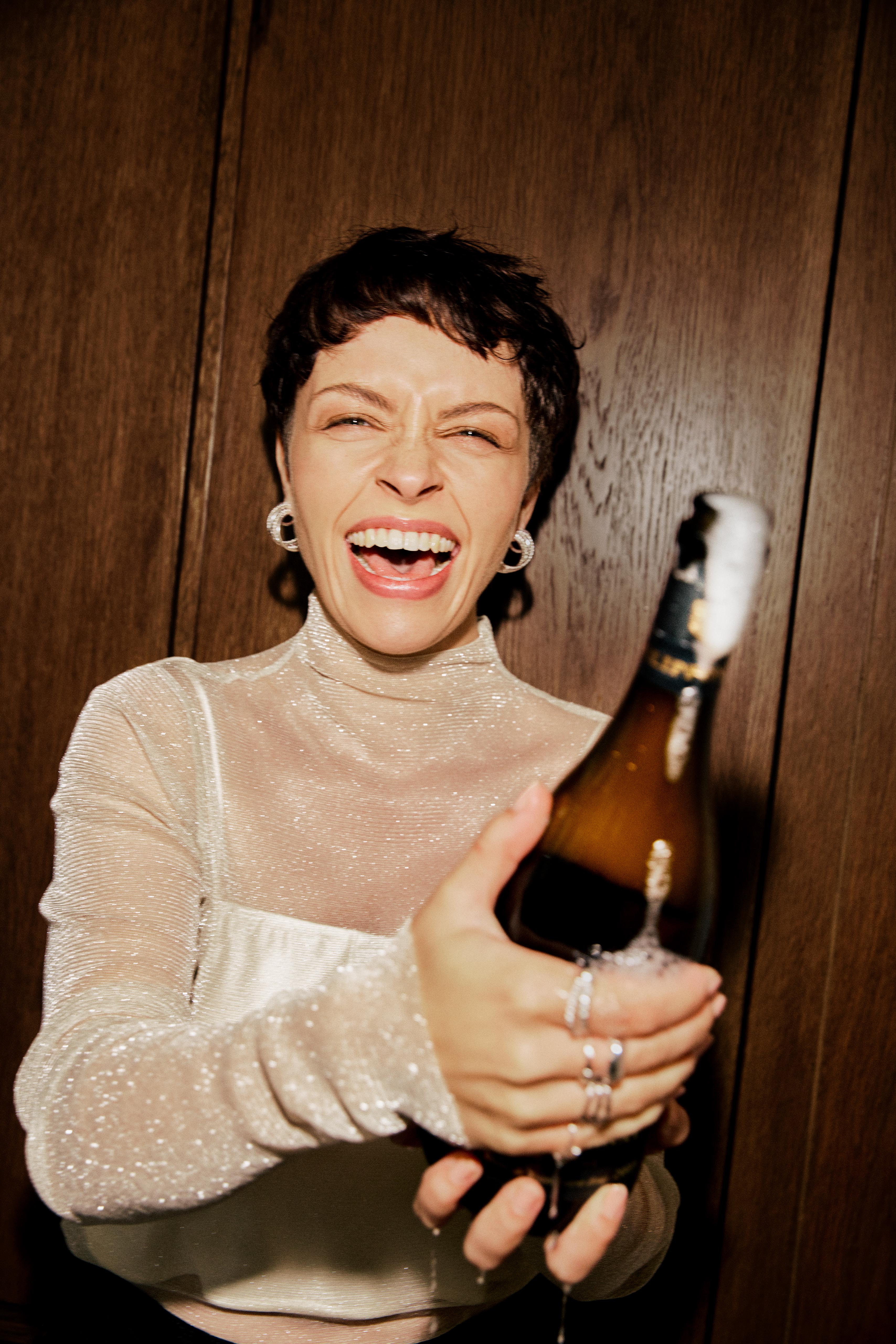 Smiling woman pops open a champagne bottle in an indoor setting with wood paneling, wearing a shimmering top and hoop earrings.