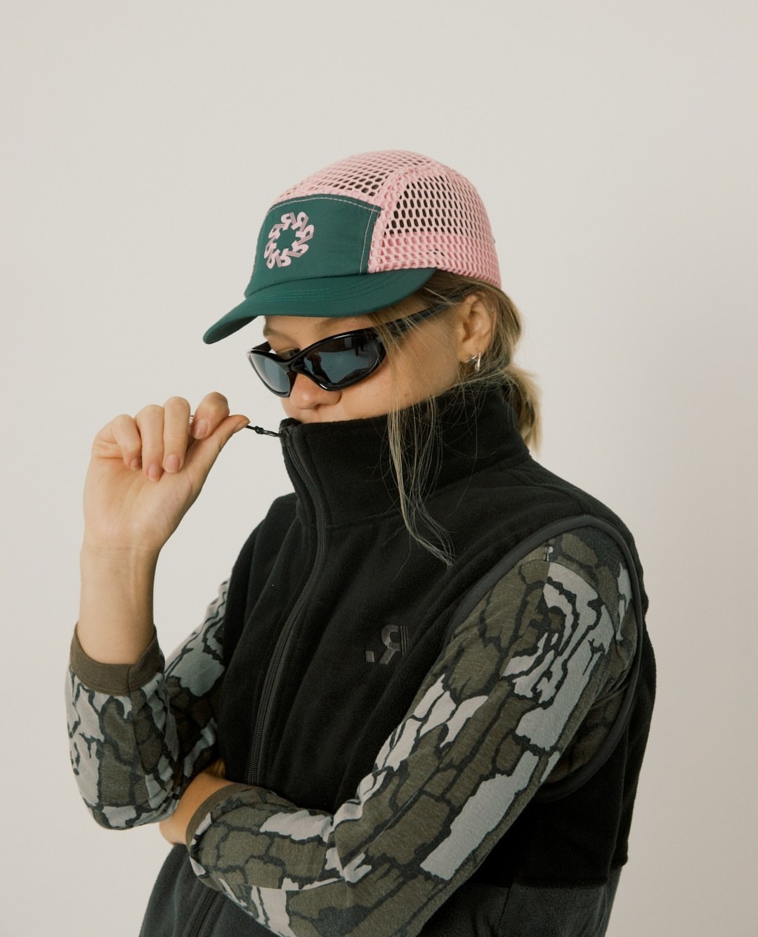 Adult woman with sunglasses poses wearing a pink and green baseball cap, black fleece vest, and printed top against a neutral background.