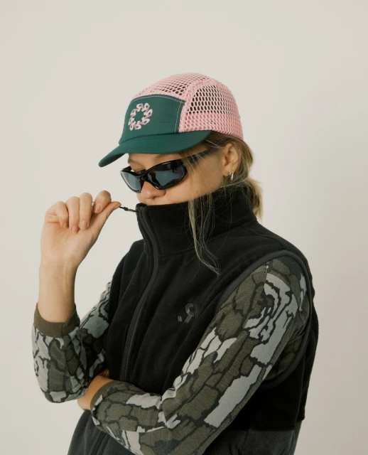 Adult woman with sunglasses poses wearing a pink and green baseball cap, black fleece vest, and printed top against a neutral background.
