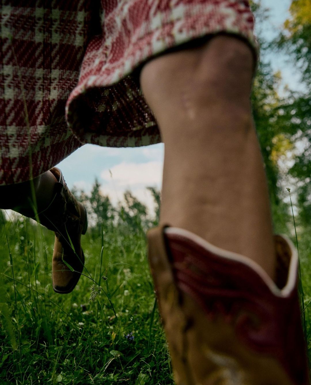 Close-up of a person's leg and foot wearing a red and white plaid skirt and brown cowboy boots, walking through a green grassy field.