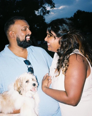 Man and woman smiling at each other while holding a small fluffy puppy outdoors at dusk