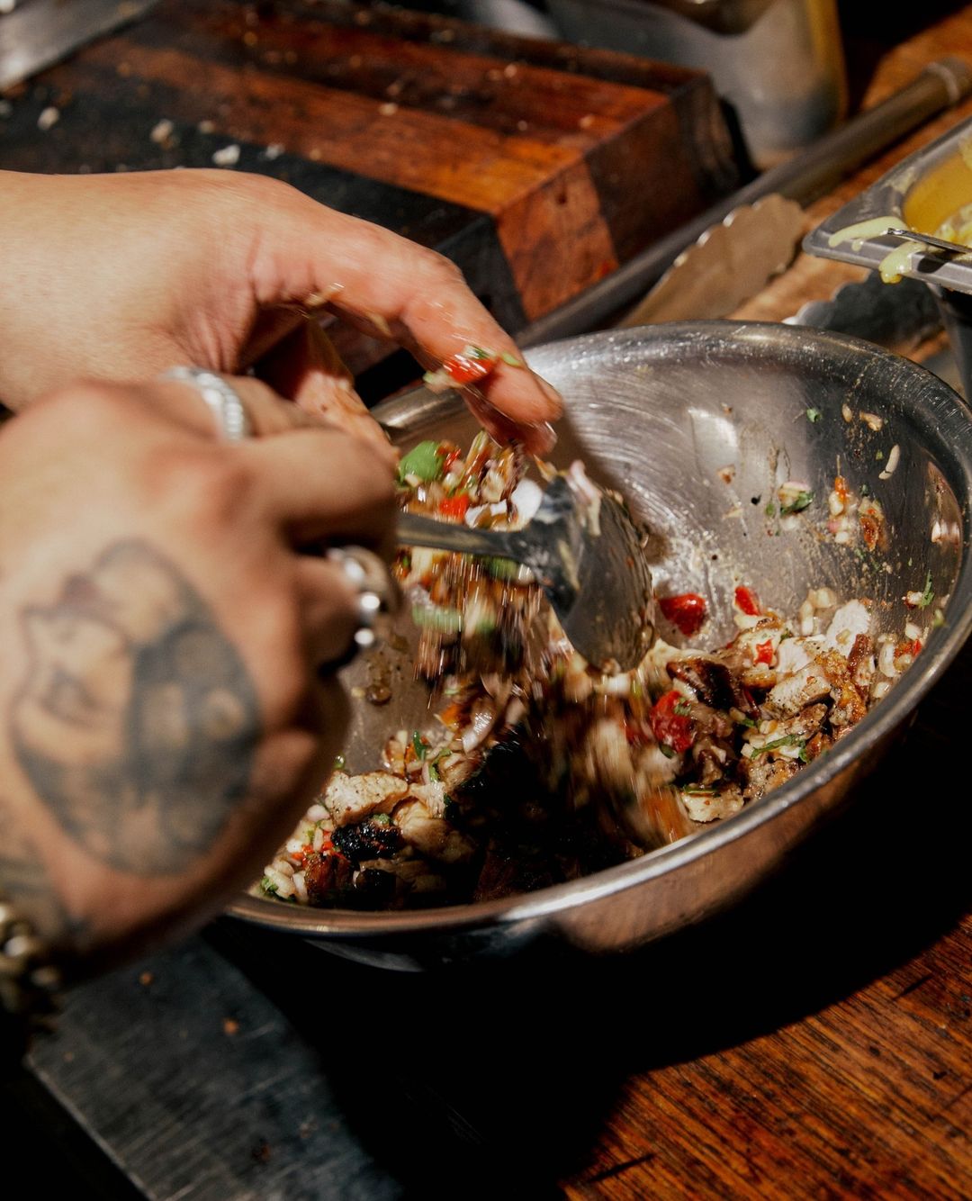 Hands toss a vibrant salad in a silver bowl, ingredients of diced vegetables and greens on a dark wood surface, close-up, eye-level angle.