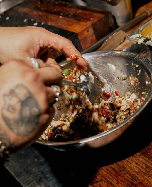 Hands toss a vibrant salad in a silver bowl, ingredients of diced vegetables and greens on a dark wood surface, close-up, eye-level angle.