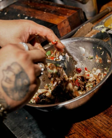 Hands toss a vibrant salad in a silver bowl, ingredients of diced vegetables and greens on a dark wood surface, close-up, eye-level angle.