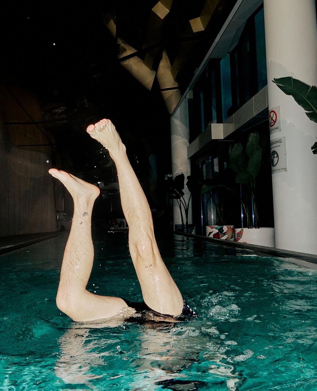 Person standing upside down in a pool with legs straight up, water ripples, and indoor pool setting with plant and dark ceiling.