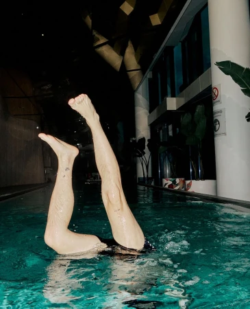 Person standing upside down in a pool with legs straight up, water ripples, and indoor pool setting with plant and dark ceiling.