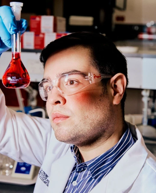 Adult male scientist wearing safety glasses and lab coat holding up a beaker with red liquid in a laboratory setting.