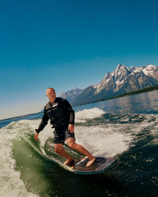 Man wake surfing on a lake with mountains in the background under a clear blue sky, wearing a black life vest and shorts, outdoor action.