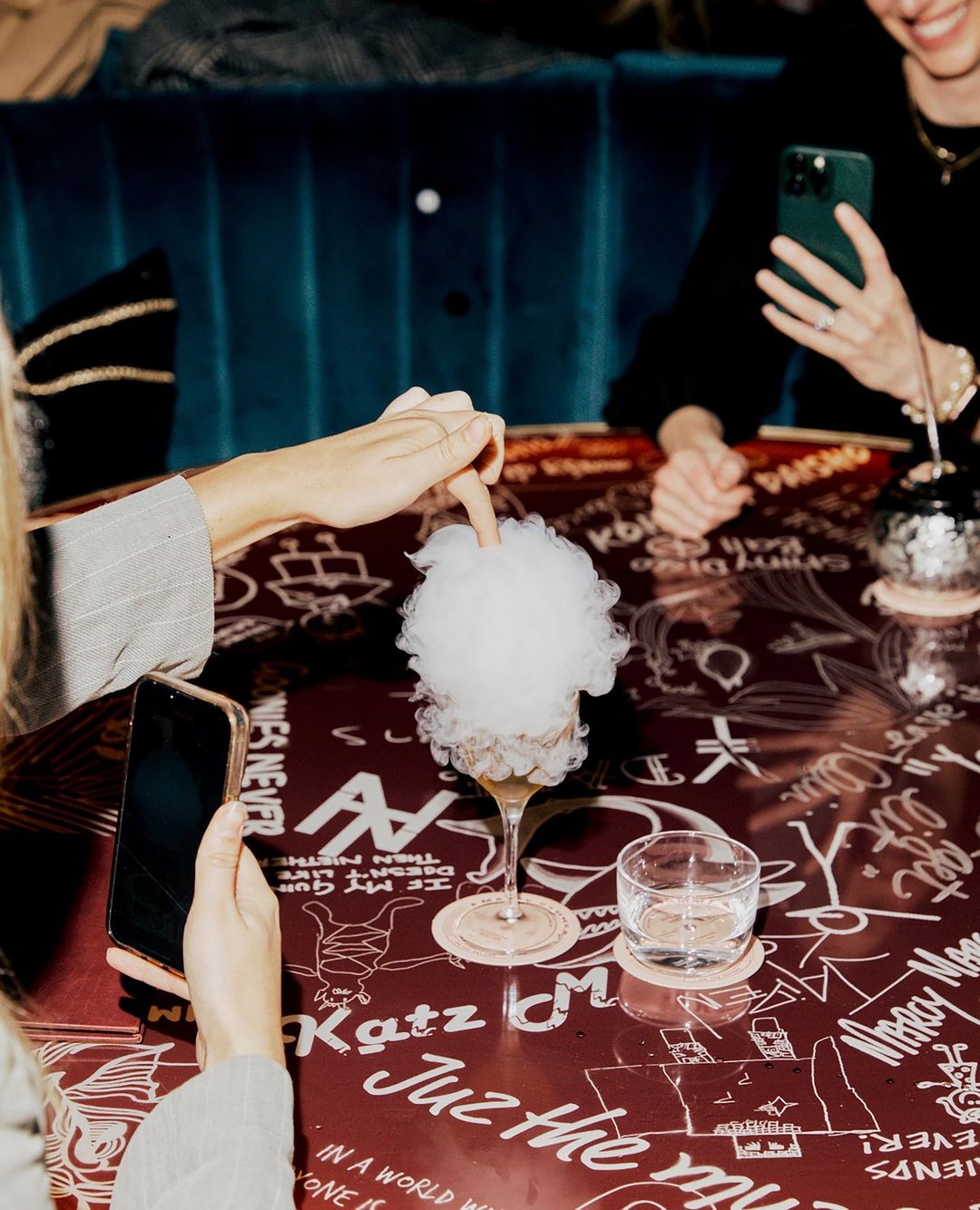 People gathered around a dark table covered in white illustrations, photographing a cocktail topped with white cotton candy.