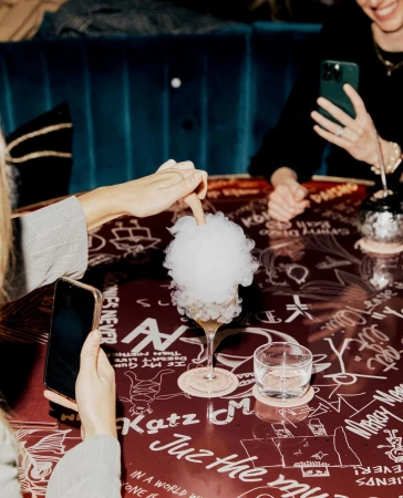 People gathered around a dark table covered in white illustrations, photographing a cocktail topped with white cotton candy.